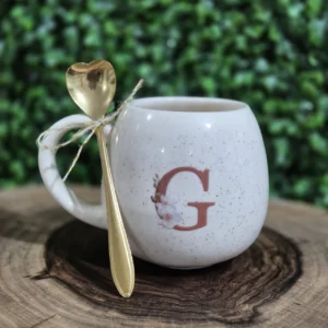 Caneca de cerâmica creme com a inicial G em arte floral e colher dourada, sobre tora de madeira