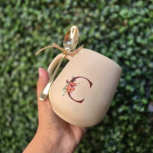 Mão segurando caneca de cerâmica creme com a inicial C em carmim e colher dourada