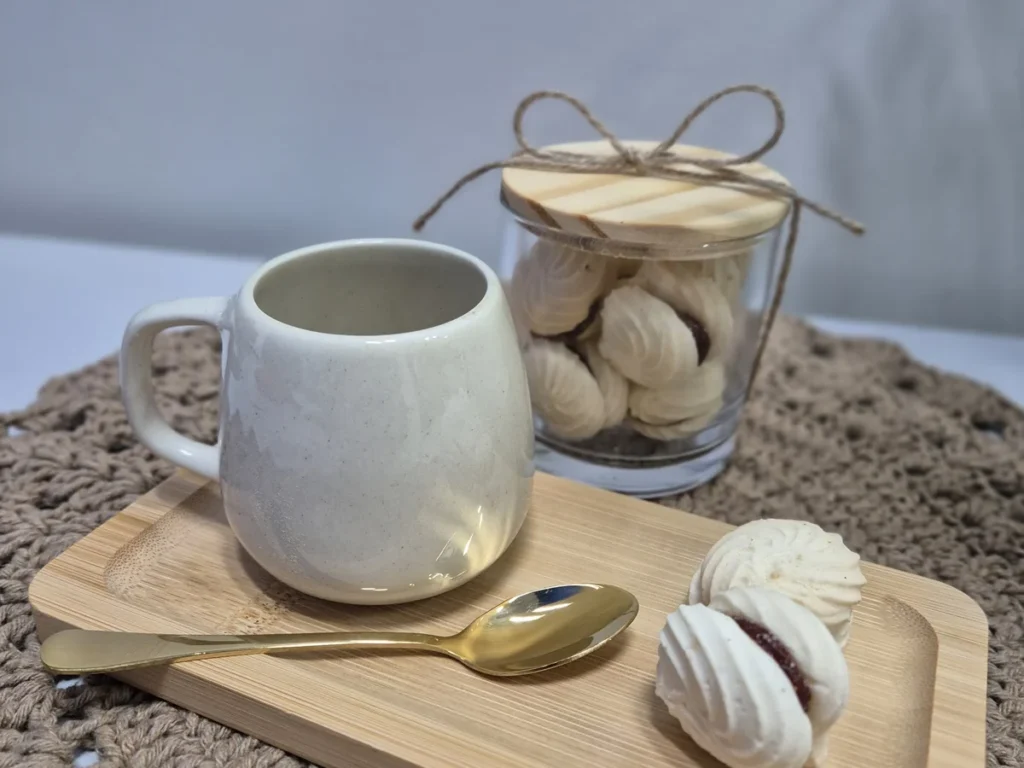 Caneca creme, colher dourada, suspiros e pote de vidro com tampa de madeira sobre tábua de bambu