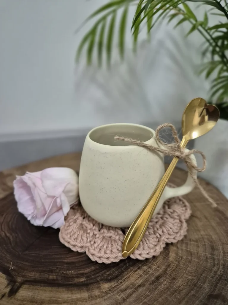 Caneca de porcelana creme com colher dourada e laço de barbante, ao lado de uma flor, sobre descanso de crochê