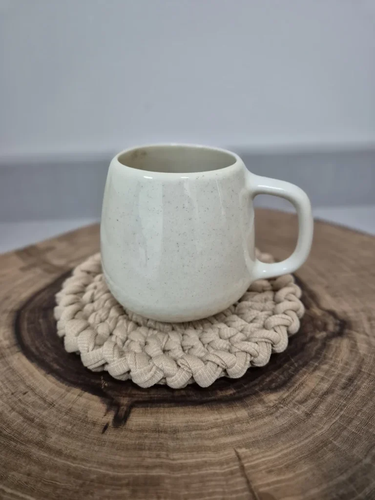 Caneca de cerâmica branca mesclada sobre descanso de copo em crochê cru, apoiada em tora de madeira