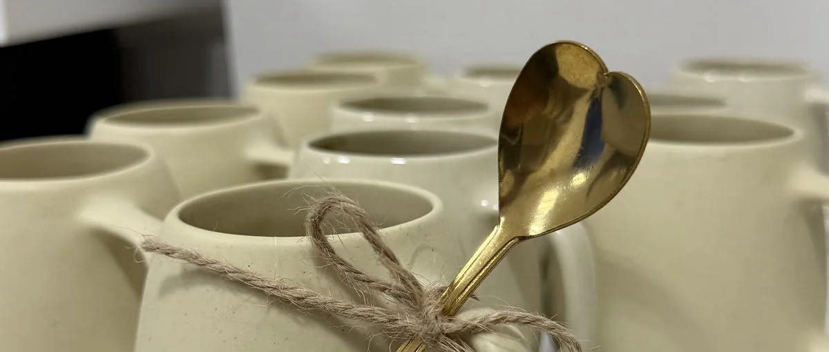 Fileiras de canecas de porcelana creme na bancada do ateliê, a da frente com colher dourada e laço de barbante