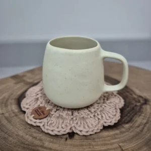 Caneca de porcelana creme sobre descanso de copo em crochê rosa, apoiada em tora de madeira