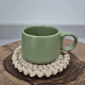 Caneca de cerâmica verde-oliva sobre descanso de copo em crochê cru, apoiada em tora de madeira