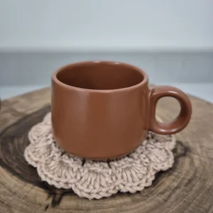 Caneca de cerâmica terracota sobre descanso de copo em crochê rosa, apoiada em tora de madeira