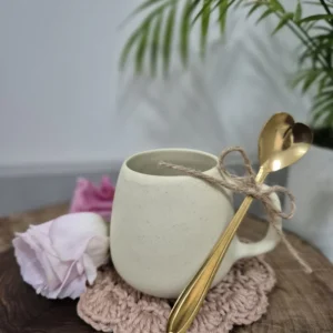 Caneca de porcelana creme com colher dourada e laço de barbante, ao lado de uma flor rosa