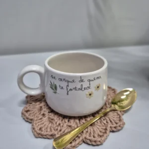 Caneca de cerâmica branca com frase escrita à mão, colher dourada e descanso de copo em crochê