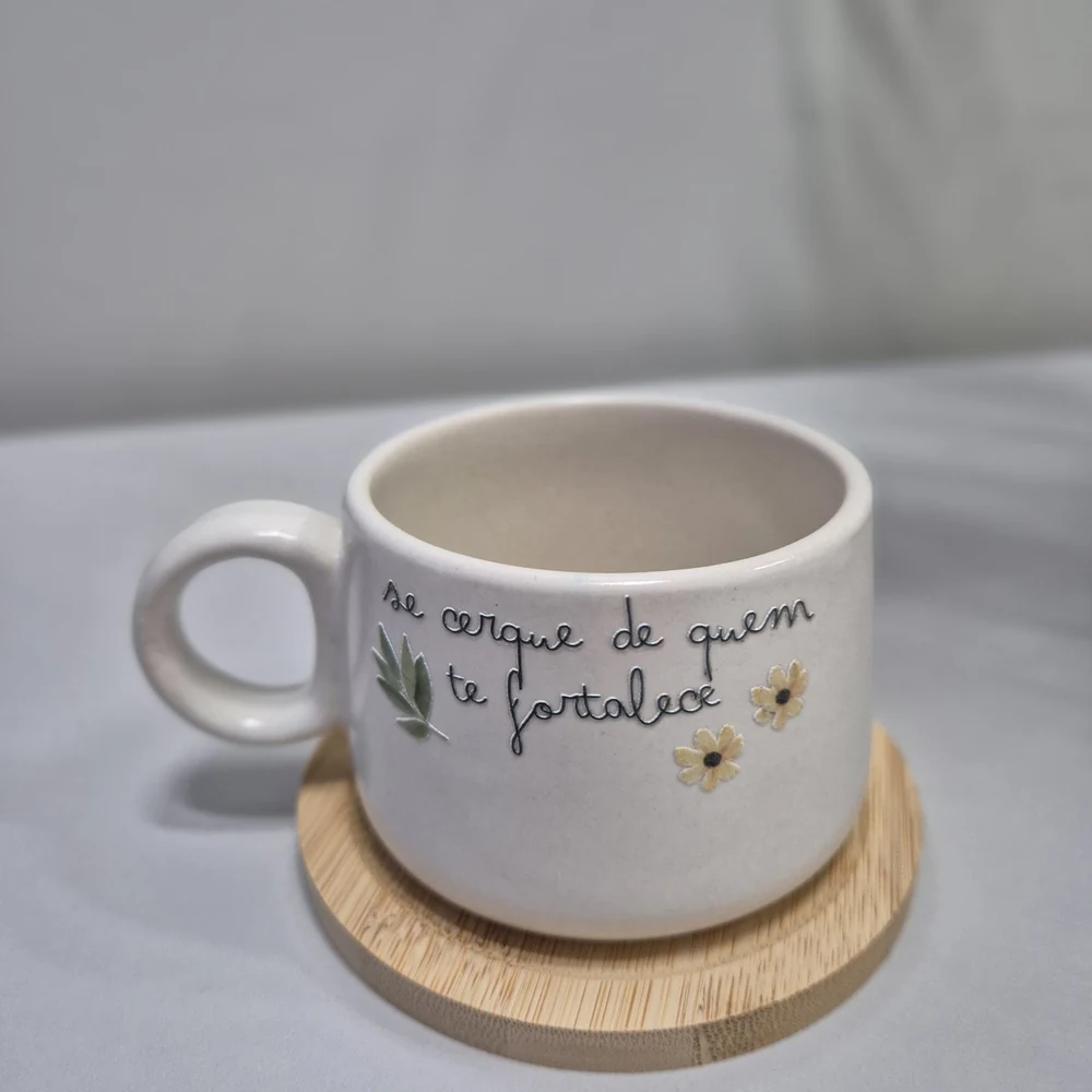 Caneca de cerâmica branca com frase escrita à mão e flores, sobre base redonda de bambu