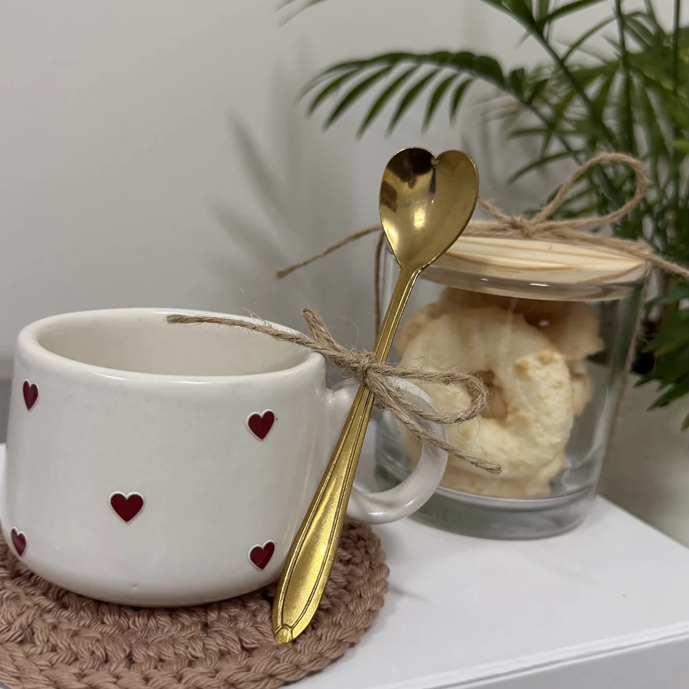 Caneca branca com corações vermelhos e colher dourada ao lado de um pote de vidro com biscoitos