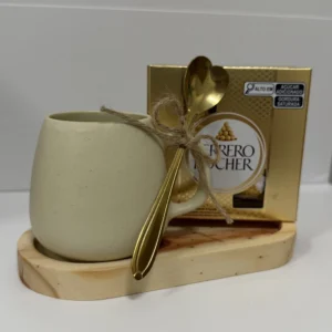 Caneca creme com colher dourada e laço de barbante ao lado de uma caixa de bombons