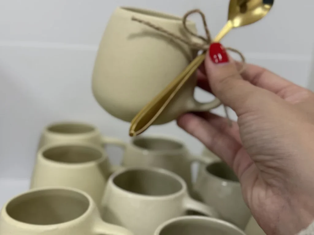 Mão segurando uma caneca de porcelana creme com colher dourada, diante de dezenas de canecas na bancada do ateliê